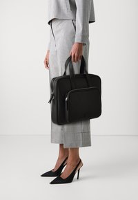 Sort læder arbejdstaske med dobbelt lynlås, to håndtag og en frontlomme, båret af en person iført et ternet outfit og sorte hæle.