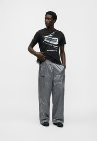 Μαύρο μπλουζάκι με γραφικό Porsche, συνδυασμένο με γκρι παντελόνι με ανακλαστικές λεπτομέρειες. Το μοντέλο κρατά μια μικρή τσάντα και φορά μαύρα παπούτσια. Μινιμαλιστικό στυλ.