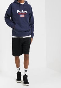 Námořnická modrá mikina s bílým logem "Dickies" a červeným textem "SINCE 1922". Obsahuje přední kapsu, kapuci se šňůrkou a černé šortky se pruhovanými ponožkami.