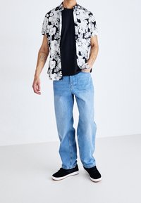 Camicia floreale bianca e nera sopra una maglietta nera, abbinata a jeans larghi blu chiaro e sneaker nere con suola bianca.