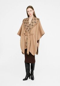Wikkel in kameelkleur met een bruin patroon op de kraag en een riem, met een losse pasvorm en brede mouwen. Draagt over een donkerbruine rok.