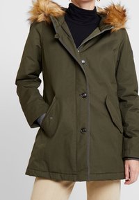 Parka en verde aceituna de longitud media con capucha de piel sintética, que cuenta con cierre de cremallera, botones a presión y bolsillos laterales. Tejido suave con textura lisa.