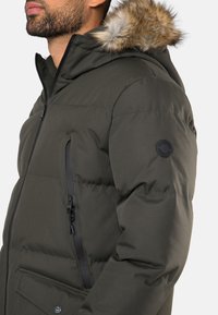 Donkergroene pufferjack met een bontgevoerd capuchon, voorzien van zwarte ritsen en een gestructureerde afwerking. Inclusief een patchdetail op de mouw.