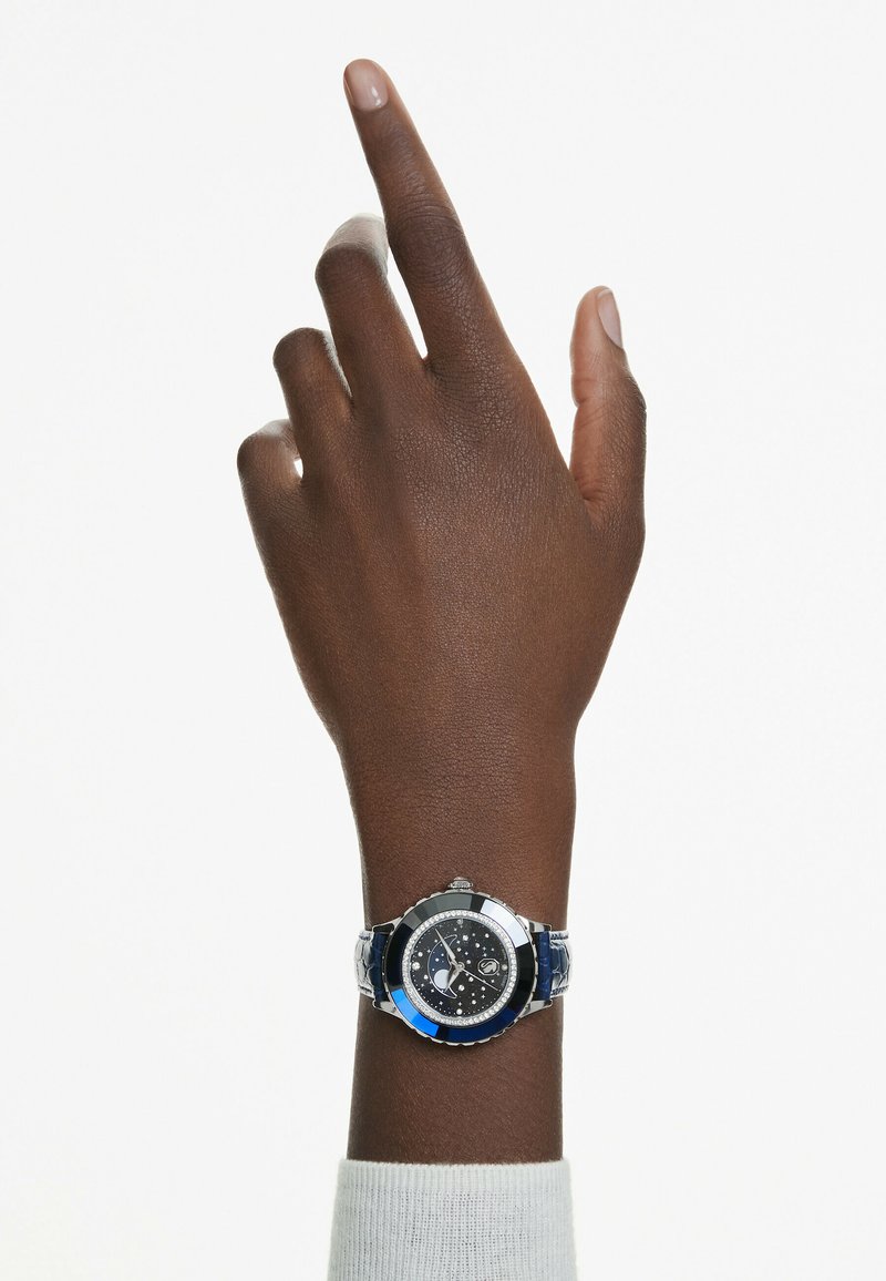 Srebrny i niebieski zegarek na nadgarstku, z ciemnym cyferblatem zdobionym diamentami, metalową ramką oraz wzorzystym skórzanym paskiem.