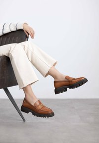 Mocasines de cuero marrón con una suela de goma negra gruesa, que presentan una pequeña tira decorativa. Llevados con pantalones cropped crema, sentados.