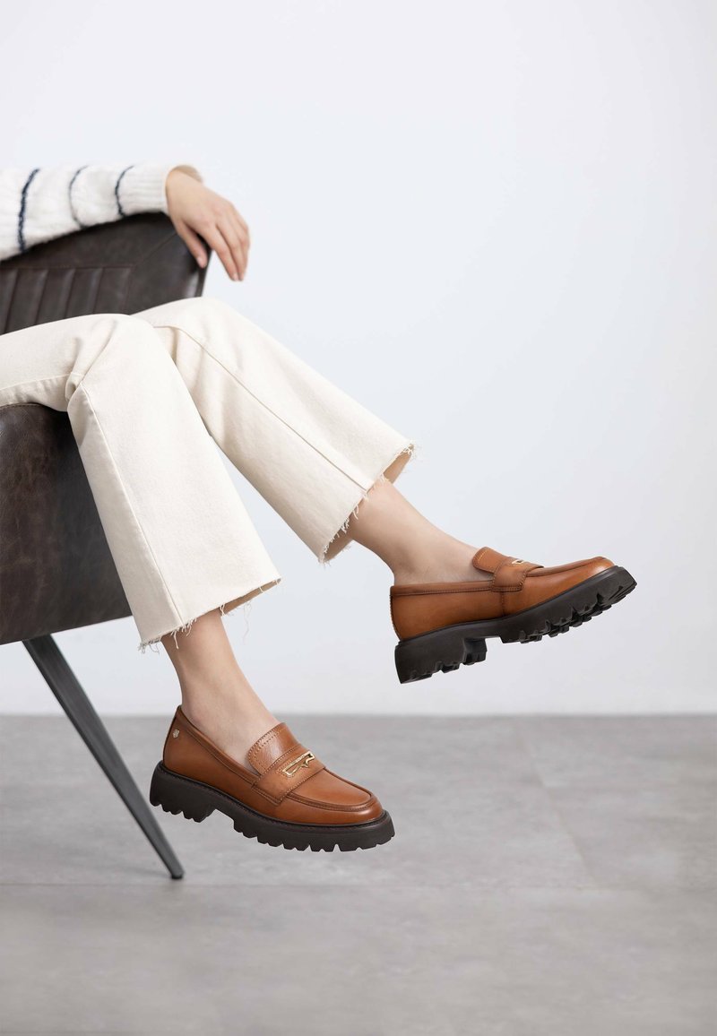 Mocasines de cuero marrón con una suela de goma negra gruesa, que presentan una pequeña tira decorativa. Llevados con pantalones cropped crema, sentados.