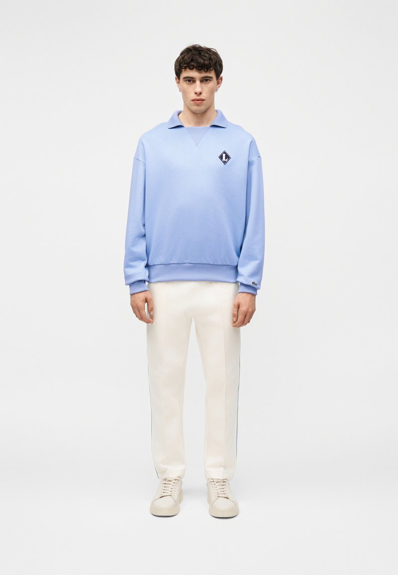 Jeune homme debout portant un sweat-shirt col roulé bleu clair à manches longues, pantalon blanc et baskets beiges sur fond blanc uni.