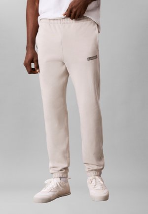 Άνδρας που φοράει μπεζ παντελόνι φόρμας Calvin Klein και λευκά αθλητικά, στέκεται μπροστά σε απλό γκρι φόντο.