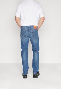 Hombre vestido con jeans rectos de color azul claro con bolsillos traseros, una camiseta blanca y zapatos negros, de pie con las manos en los bolsillos traseros.