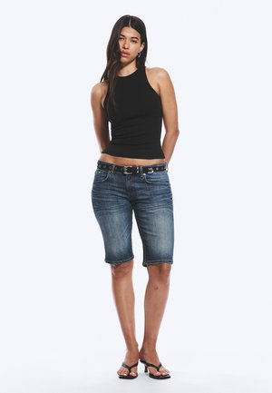 Woman with long dark hair wearing a sleeveless black top, knee-length denim shorts with a belt, and black heeled sandals, standing with hands behind back.
