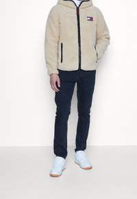 Giacca in pile beige con zip e rifiniture nere, jeans slim-fit scuri e sneakers bianche con suole color gomma su una persona in piedi.