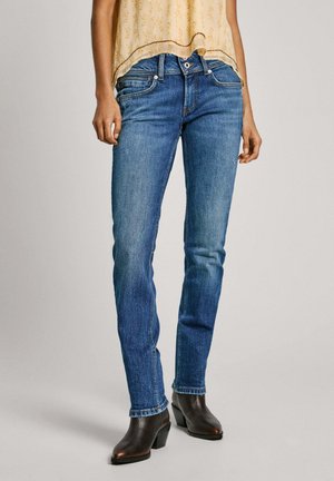 Woman wearing medium-wash slim-fit jeans, light yellow patterned blouse, and dark brown leather heeled ankle boots, standing on plain background.