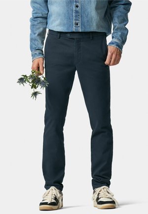 Navy blue straight-leg pants featuring a smooth texture and front pockets, paired with black and beige sneakers. A hand holds green foliage.