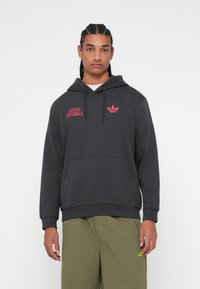Čierna mikina s kapucňou so sladkým nápisom "ADIDAS ORIGINALS" a logom v červenej farbe. Obsahuje predné vrecko a kapucňu s šnúrkou. Hladká textúra, voľný strih.
