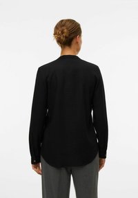 Chemise noire à manches longues en tissu lisse, avec un ourlet incurvé et des poignets boutonnés, vue de derrière.