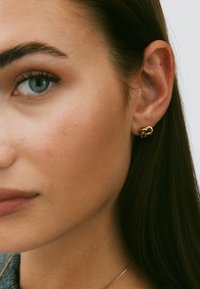 Kullast nööp-kõrvarõngas sõlmitud disainiga. Sile tekstuur ja läikiv viimistlus. Kõrvalvaade kõrvarõngast mudeli kõrvas.