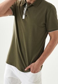 Polo shirt verde oliva realizzato in tessuto texture, con colletto classico, pattina con bottoni e dettaglio logo bianco sul colletto.