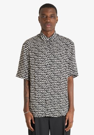 Persona in piedi rivolta in avanti, che indossa una camicia bianca e nera a maniche corte con motivo a zigzag e pantaloni neri su sfondo semplice.