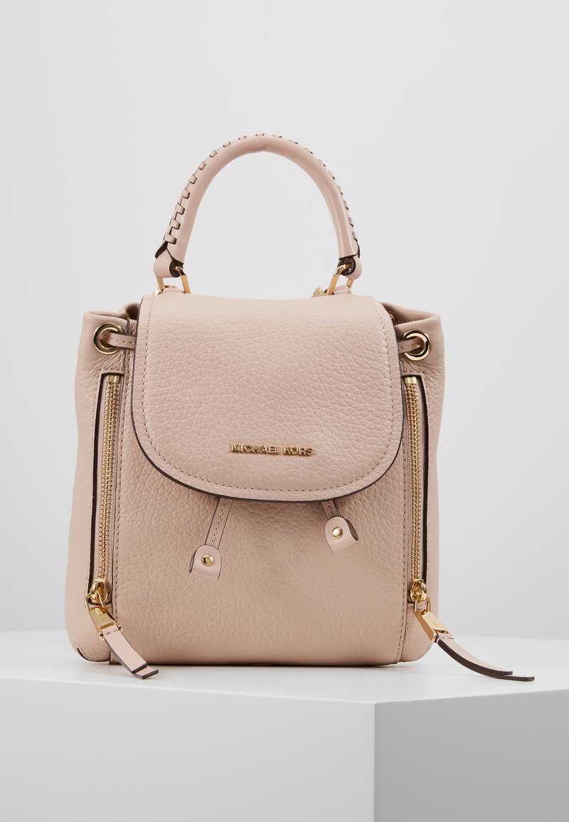 Sac à dos en cuir rose clair avec rabat, ferrures dorées, deux poches zippées et finition texturée. Il dispose d'une poignée supérieure et de bretelles ajustables.