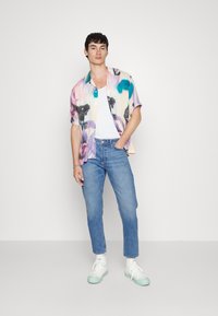 Camisa de manga curta, com botão, em padrão de aquarela pastel; combinada com jeans azul claro e ténis brancos. Look simples e casual.