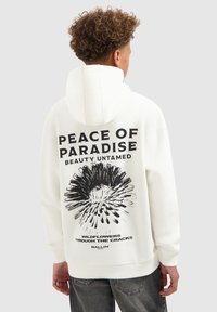 Jongere met krullend haar draagt een witte hoodie met een zwart bloemenafbeelding en de tekst "Vrede van het Paradijs, Ongerepte Schoonheid, Wilde Bloemen door de Scheuren."