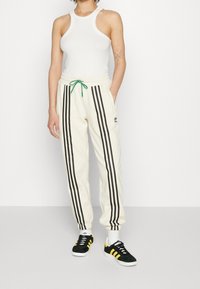 Pantaloni jogger crema con tre strisce verticali nere, cintura elastica con coulisse verde e tasche laterali. Indossati con sneakers nere a bassa trama.