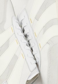 Baskets blanches en matière synthétique texturée, avec des accents en daim, des lacets blancs plats et un design rayé dans une palette de couleurs claires.