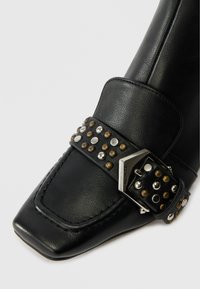 Black leather shoe with a square toe, featuring a studded strap and a metallic buckle. The strap has round studs in silver and bronze.