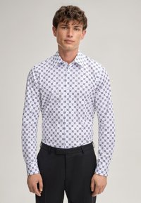 Chemise blanche avec un motif géométrique répétitif en bleu marine, col pointu, fermetures à boutons et manches longues, associée à un pantalon noir.