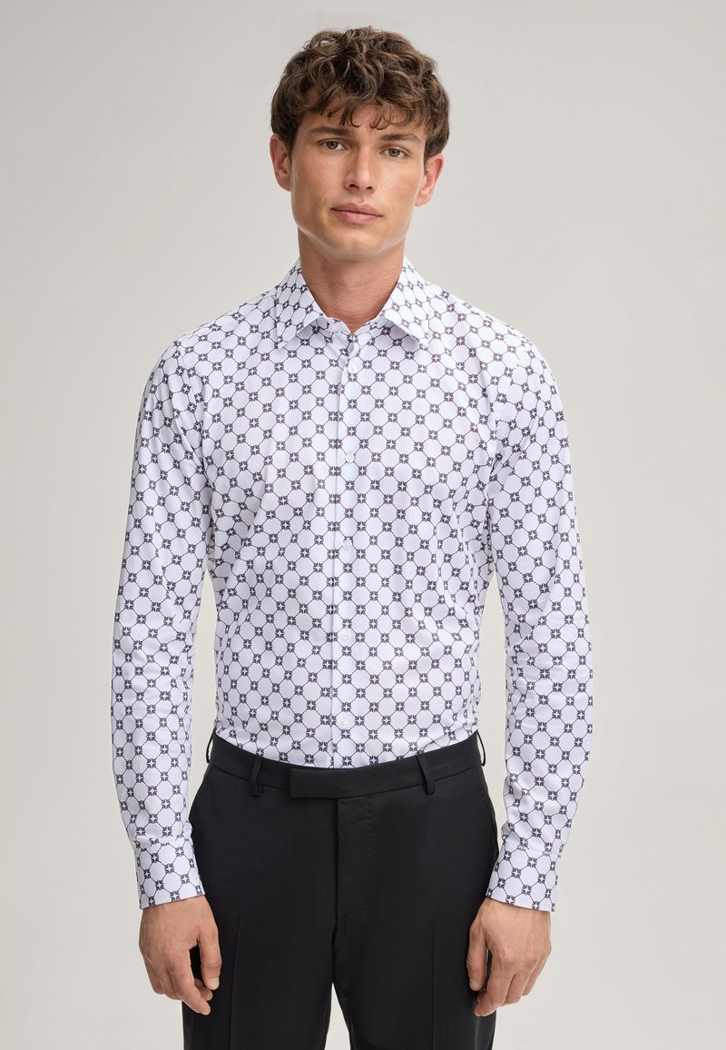 Chemise blanche avec un motif géométrique répétitif en bleu marine, col pointu, fermetures à boutons et manches longues, associée à un pantalon noir.