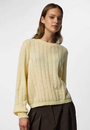 Jeune femme portant un pull jaune clair en maille et un pantalon plissé marron foncé sur un fond blanc uni.