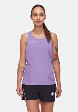 Kobieta z brązowym kucykiem, ubrana w fioletowy, bezrękawnik Mammut oraz czarne spodenki Mammut, prezentująca kwiatowe i sercowe tatuaże na ramionach.