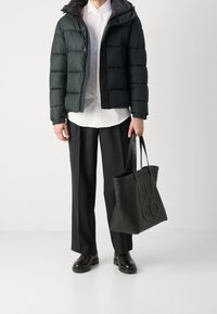 Uomo che indossa una giacca a vento nera, una camicia bianca, pantaloni neri e stivali neri, mentre tiene in mano una grande borsa tote nera con "HUGO" impresso sul davanti.