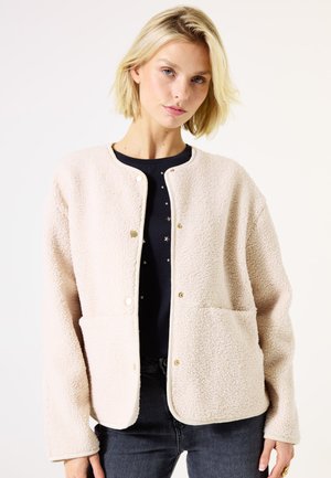 Veste en polaire beige avec une surface texturée, un col rond, deux poches avant, des boutons-pressions et un liseré blanc contrastant.
