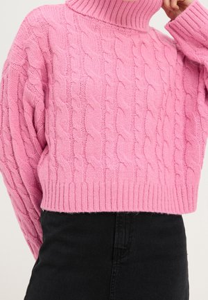 Personne portant un pull à col roulé en maille torsadée rose et un pantalon noir, avec une main touchant le col du cou.