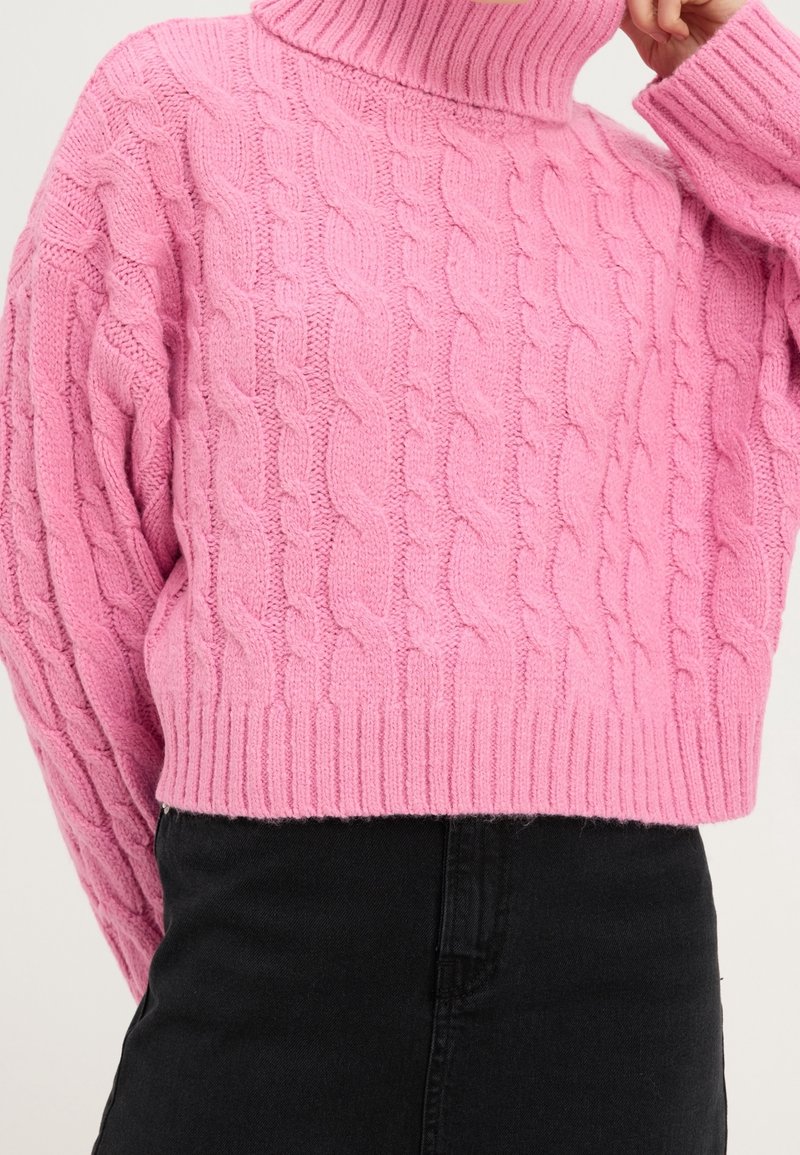 Personne portant un pull à col roulé en maille torsadée rose et un pantalon noir, avec une main touchant le col du cou.