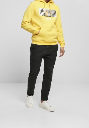 Persona che indossa una felpa gialla con stampa grafica, pantaloni neri e sneakers bianche, in piedi con una mano in tasca.