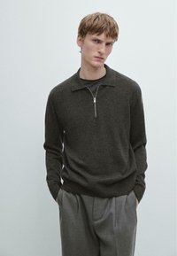 Maglione con zip grigio scuro con colletto e maniche lunghe, realizzato in materiale morbido. Indossato sopra una camicia nera, abbinato a pantaloni grigi.