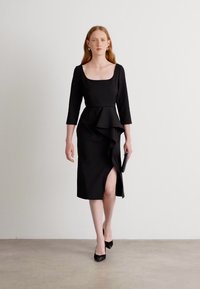 Abito midi nero con scollo a quadrato, maniche a tre quarti, dettagli a rouches sovrapposti e uno spacco laterale, abbinato a una pochette nera.