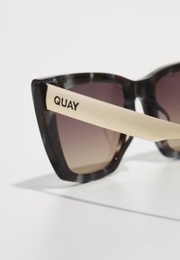 Kattenoog zonnebril met een donker schildpadframe, gradient bruine lenzen en een arm met gouden accenten met de merknaam "QUAY."