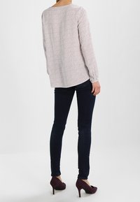 Langärmlige, weiße Bluse mit einem kleinen roten Muster. Getragen mit dunkelblauen Slim-Fit-Jeans und burgunderfarbenen High Heels. Glatter Stoff und einfaches Design.