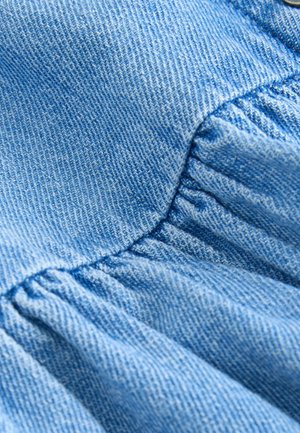 Hellblauer Denimstoff mit strukturierter Oberfläche, der entlang der Naht geraffte Falten aufweist. Das Material zeigt ein feines Webmuster.