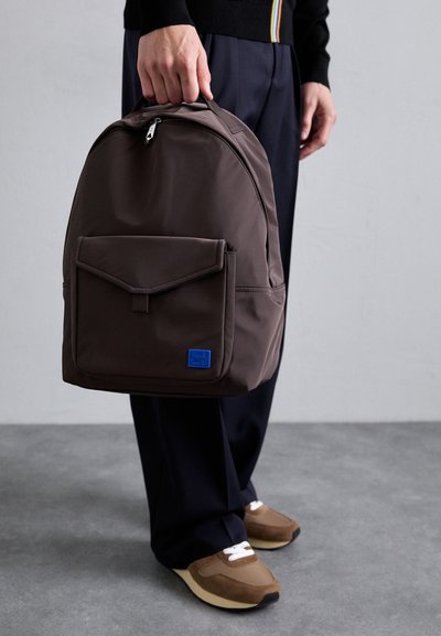 Mochila de tela marrón con un compartimento principal con cremallera, bolsillo en la solapa frontal y logotipo azul. Textura suave, forma redondeada y asas acolchadas.