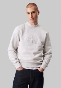 Sudadera gris claro con cuello redondo acanalado, que presenta un logo "CK" en relieve. Combinada con unos vaqueros oscuros de corte relajado, con las manos en los bolsillos.