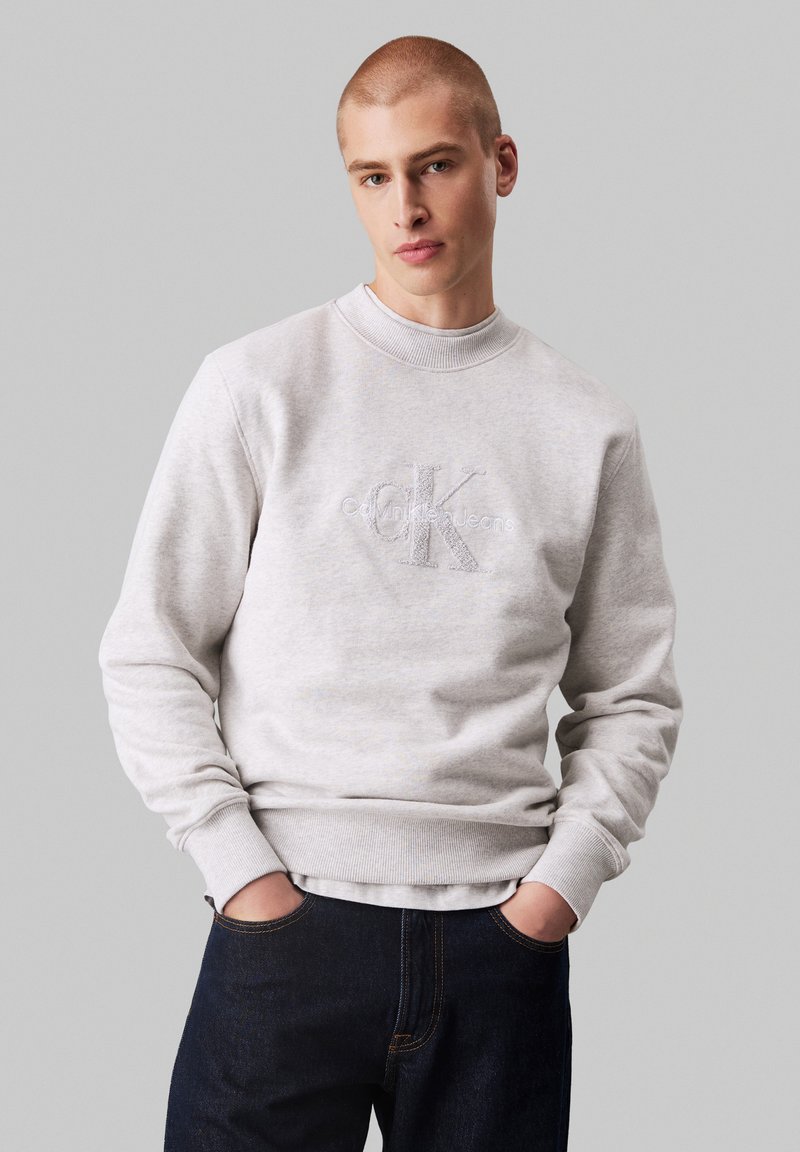 Sudadera gris claro con cuello redondo acanalado, que presenta un logo "CK" en relieve. Combinada con unos vaqueros oscuros de corte relajado, con las manos en los bolsillos.