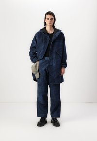 Jūras flotes teksturēta jaka ar kapuci, saskaņota ar atbilstošām biksēm. Abiem ir raksts, bet rokā turētā pelēkā cepure. Melna apavi pabeidz izskatu.