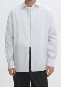 Chemise rayée blanche avec un design à boutons, manches longues et col classique. Fabriquée en tissu léger à texture lisse.