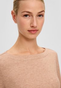 Close-up van een persoon die een lichtbruine top met een zachte textuur draagt, voorzien van een brede halslijn en een ontspannen pasvorm, met minimale stikdetails.