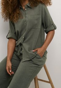 Donna con capelli ricci che indossa una camicia a maniche arrotolate di colore verde oliva e pantaloni abbinati, seduta su uno sgabello di legno con una mano in tasca.