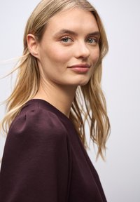 Vaalea nainen, jolla on vaalea iho ja vihreät silmät, pukeutuneena tumman violetiseen puseroon, katsomassa hiukan olkansa yli tasaisen taustan edessä.